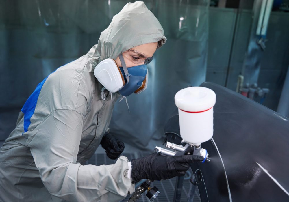 Lady automotive engineer wearing protective equipment working on car body element in paint chamber